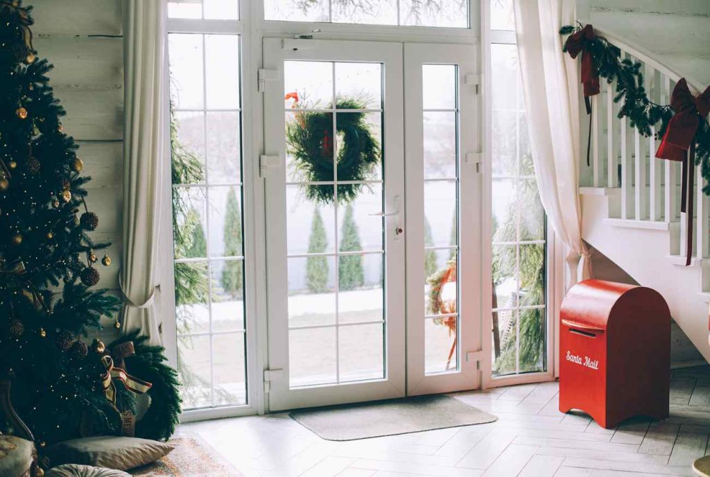 Intérieur d'une maison décorée pour Noël avec une grande porte d'entrée menuiserie pvc vitrée moderne donnant sur un jardin enneigé.